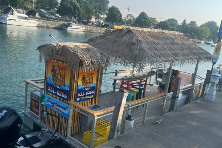 Floating tiki bar docked on a calm river with boats and trees in the background.